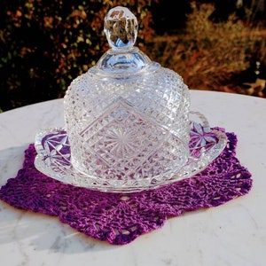 May include: A clear, diamond-cut glass butter dish with a domed lid sits on a decorative plate. The dish rests on a purple crocheted doily. The background is blurred with natural light.