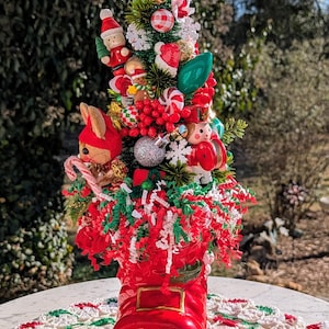 May include: A miniature Christmas tree decorated with red and green ornaments, candy canes, and a small wooden Santa figure. The tree is planted in a red boot.