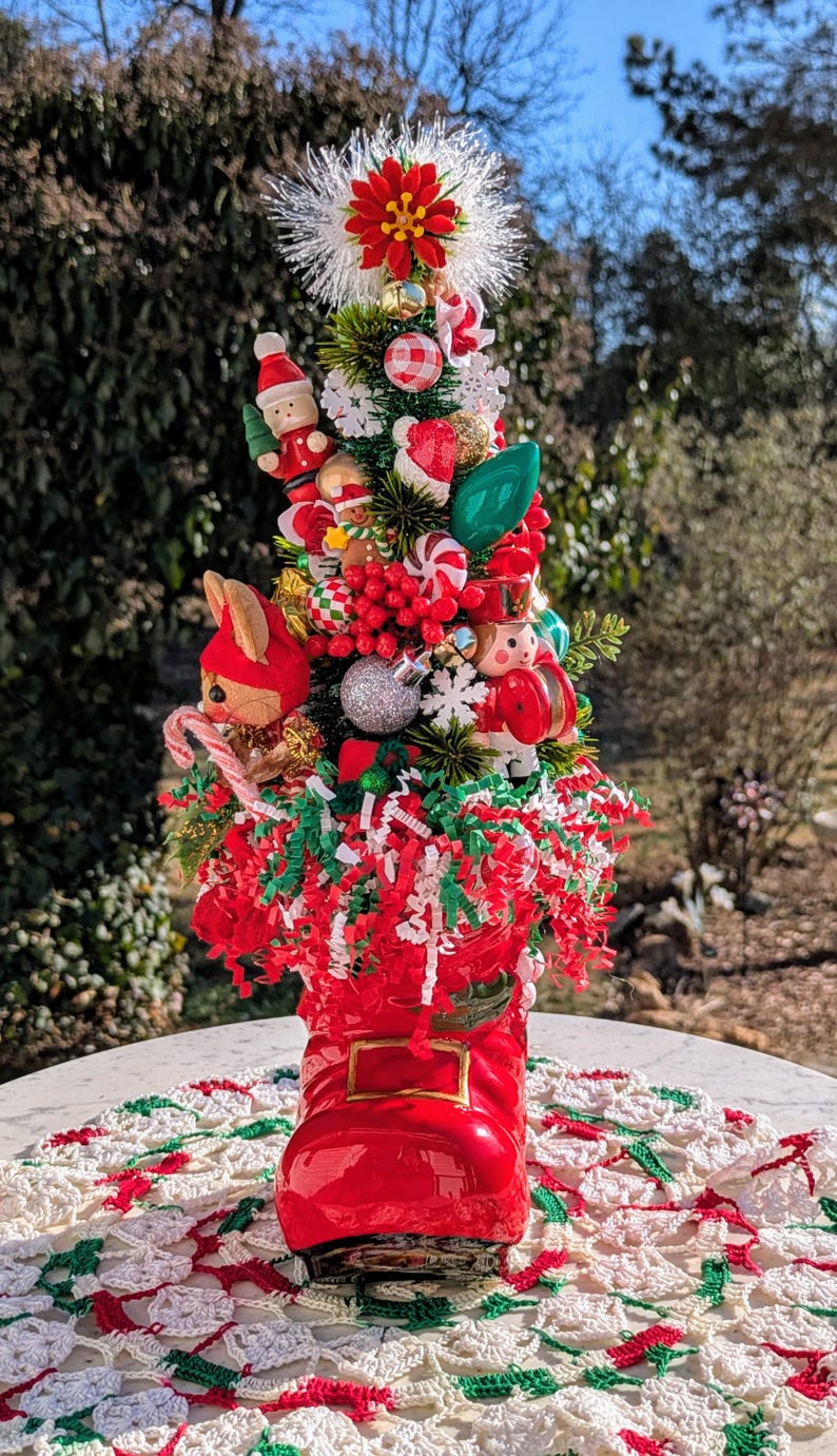May include: A miniature Christmas tree decorated with red and green ornaments, candy canes, and a small wooden Santa figure. The tree is planted in a red boot.