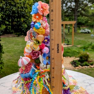 以下が含まれることがあります： パステルカラーの花、リボン、イースターエッグで飾られた、バスケットベースのカラフルなイースターツリーの装飾。ツリーの高さは約28cmです。