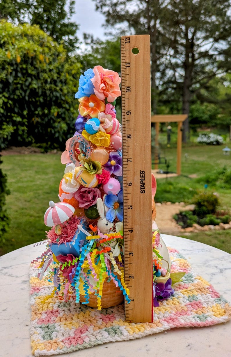 以下が含まれることがあります： パステルカラーの花、リボン、イースターエッグで飾られた、バスケットベースのカラフルなイースターツリーの装飾。ツリーの高さは約28cmです。