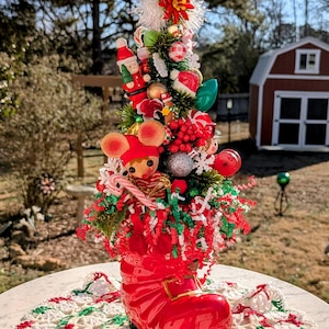 May include: A miniature Christmas tree decorated with red, green, and white ornaments, candy canes, and a small mouse figurine. The tree is planted in a red boot.