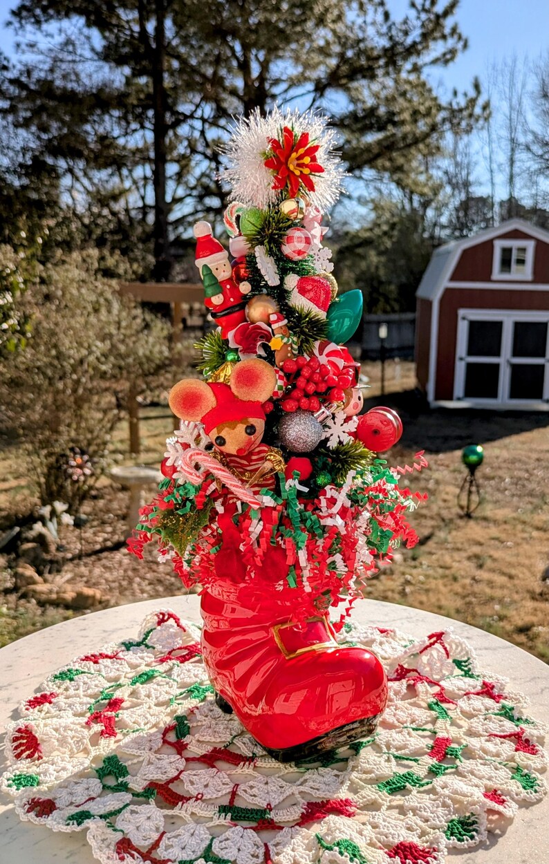May include: A miniature Christmas tree decorated with red, green, and white ornaments, candy canes, and a small mouse figurine. The tree is planted in a red boot.