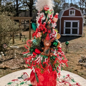 May include: A miniature Christmas tree decorated with red and green ornaments, candy canes, and a small teddy bear wearing a Santa hat. The tree is in a red boot-shaped planter.
