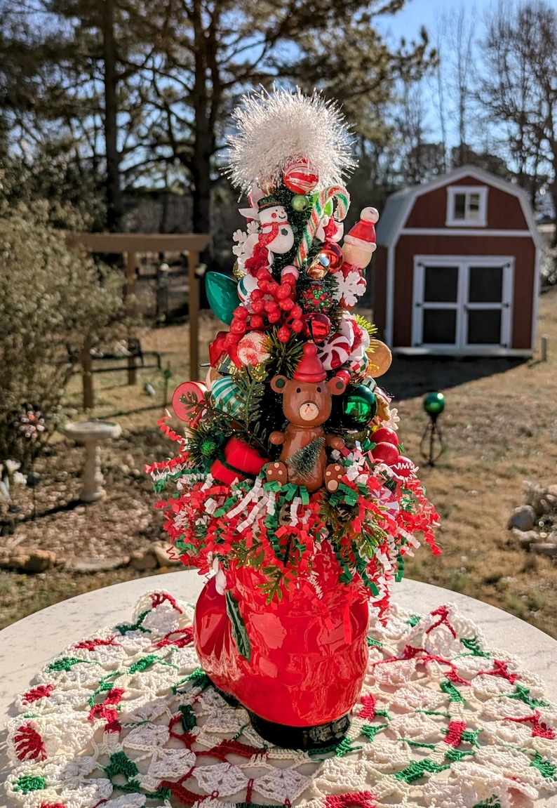 May include: A miniature Christmas tree decorated with red and green ornaments, candy canes, and a small teddy bear wearing a Santa hat. The tree is in a red boot-shaped planter.
