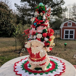 May include: A vintage Santa Claus figurine holding a Christmas tree decorated with ornaments, candy canes, and a "Merry Christmas" banner. The tree is on a white crocheted doily with red, green, and white stripes.