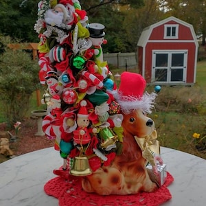 May include: A whimsical Christmas tree decorated with ornaments, ribbons, and miniature figures, including Santa Claus and snowmen. A ceramic dog wearing a Santa hat sits at the base, on a red crocheted doily. The tree is approximately 18 inches tall.