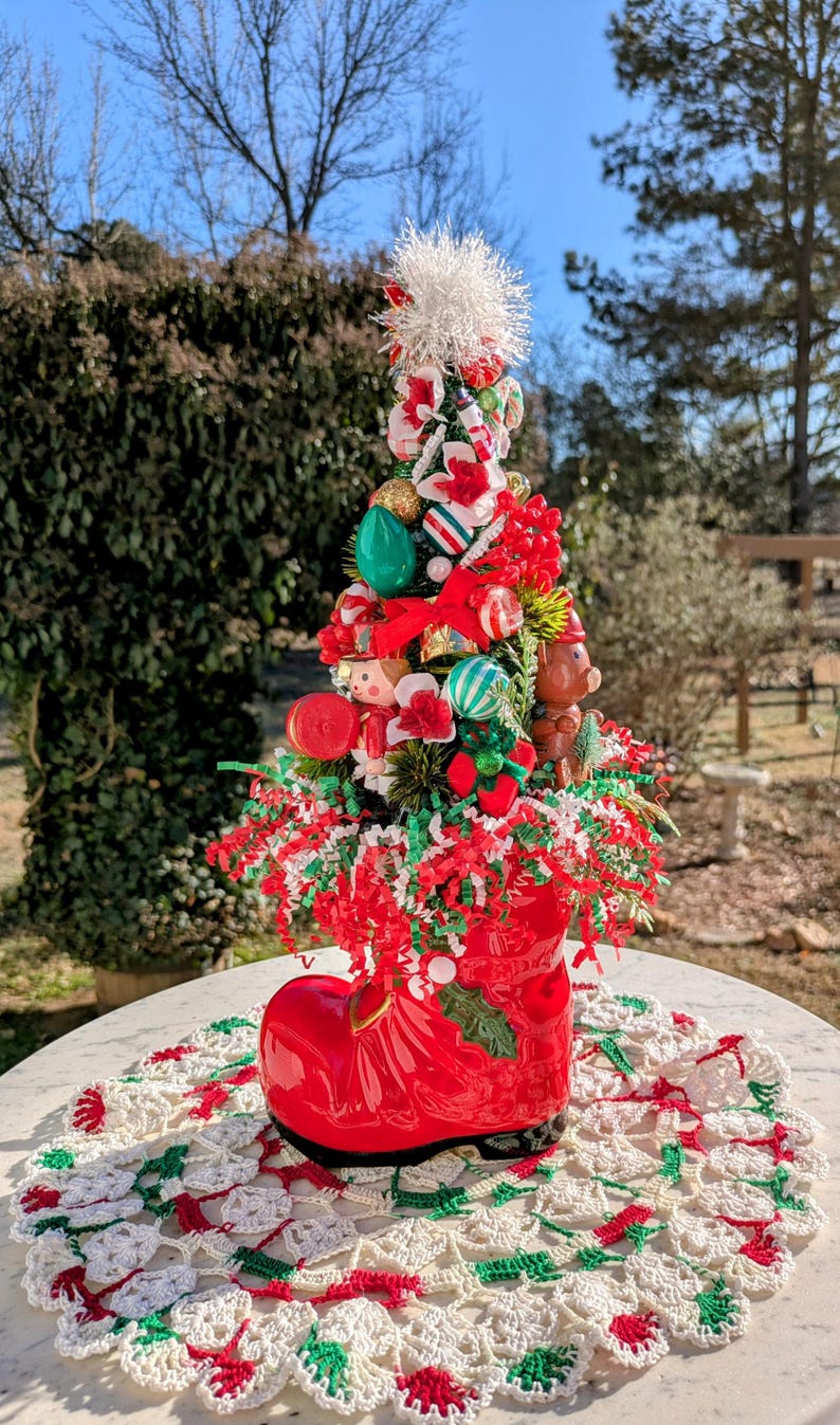 May include: A miniature Christmas tree decorated with ornaments, candy canes, and a small toy soldier, all nestled in a red boot planter. The boot sits on a white crocheted doily.
