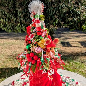 May include: A red boot-shaped planter with a miniature Christmas tree inside. The tree is decorated with red and green ornaments, a white pom-pom, and a small mouse figurine. The planter is sitting on a white crocheted doily.