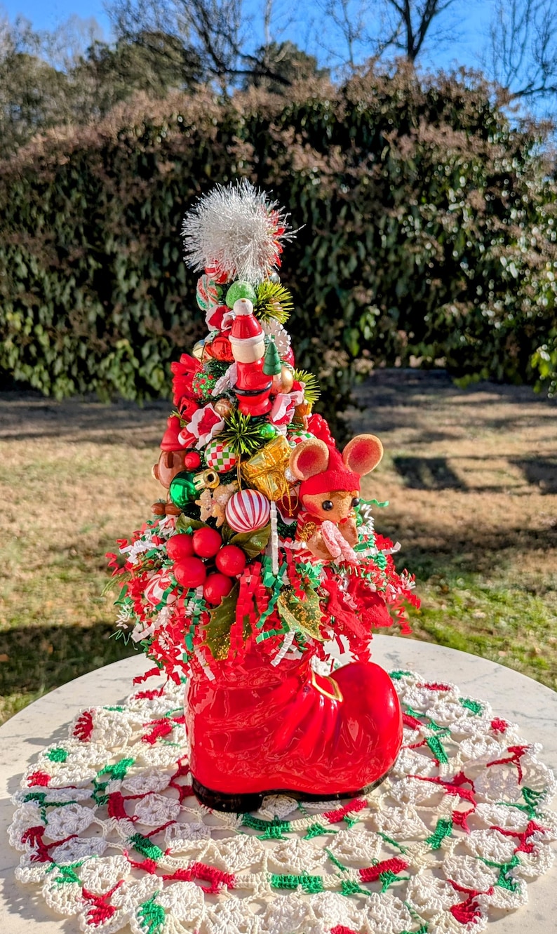 May include: A red boot-shaped planter with a miniature Christmas tree inside. The tree is decorated with red and green ornaments, a white pom-pom, and a small mouse figurine. The planter is sitting on a white crocheted doily.