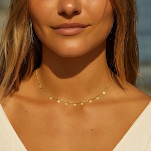 Peut inclure: Un délicat collier en or avec de petits pendentifs ronds espacés le long de la chaîne. Le collier est porté autour du cou, créant un look subtil et élégant. La couleur or offre une lueur chaude et rayonnante.