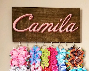 May include: A wooden sign with the name "Camila" in pink letters, with hooks for hanging hair bows. The hooks are filled with colorful hair bows in various patterns and colors.