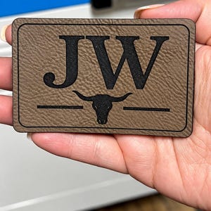 May include: A rectangular brown patch with a textured surface featuring the black engraved letters JW above a black graphic of a longhorn bull skull. The patch has a thin black border line around the entire perimeter of the rectangular shape.