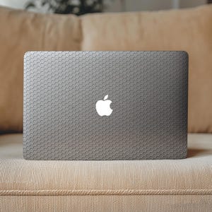 May include: A dark gray laptop with a repeating hexagonal honeycomb pattern covering the entire outer lid surface. The iconic white Apple logo is centered on the lid, contrasting against the dark textured geometric design of the laptop skin.