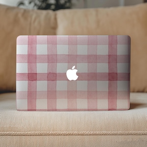 May include: A laptop computer with a pink and white watercolor gingham pattern. The Apple logo is visible in white. The laptop is resting on a beige couch.