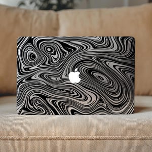 May include: Laptop with a black and white swirling pattern. The design features a fluid, marbled effect with a glossy finish. The Apple logo is visible in white. The laptop is resting on a beige sofa.