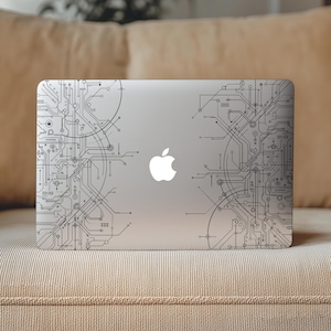 Może przedstawiać: Srebrny laptop z czarno-białym wzorem płytki drukowanej na pokrywie. Wzór zawiera szczegółowe linie i kształty, z logo Apple pośrodku. Laptop spoczywa na beżowej powierzchni.