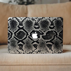 Puede incluir: Una pegatina para portátil con un patrón de piel de serpiente en blanco y negro de alto contraste que cubre toda la superficie exterior. El diseño muestra una textura de escamas repetitiva con un logotipo de Apple blanco centrado.