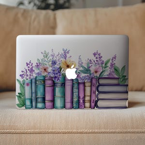 Puede incluir: Un ordenador portátil blanco con un diseño de libros y flores. El diseño presenta una fila de libros coloridos en tonos azules, morados y turquesas, coronados por flores de acuarela en morado, rosa y amarillo. El logotipo de Apple es visible.