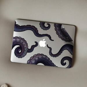 Op de afbeelding: Een laptop met een decoratieve skin met een paars en blauw octopus tentakel ontwerp. De tentakels zijn over het zilveren oppervlak gerangschikt, met het Apple-logo in het midden.