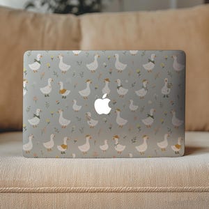 May include: A laptop skin featuring a repeating pattern of white geese on a light gray background. The geese are depicted in various poses, some wearing small hats, scarves, or floral wreaths, interspersed with tiny yellow and orange flowers.