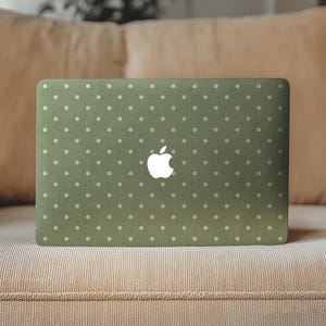 May include: A laptop with a matte olive green skin featuring a repeating pattern of small, light-colored polka dots. The center of the laptop lid displays a white Apple logo. The surface of the skin has a subtle, textured appearance.