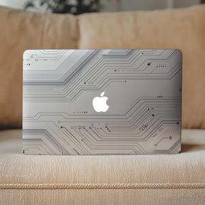 Op de afbeelding: Een zilveren laptop met een geprint printplaatpatroon op de deksel. Het ontwerp bestaat uit ingewikkelde zwarte lijnen, stippen en geometrische vormen tegen een lichtgrijze achtergrond, met een oplichtend wit Apple-logo in het midden.