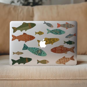 May include: A laptop covered in colorful fish stickers. The fish designs vary in size, color, and pattern, including shades of green, orange, blue, and yellow. The laptop is resting on a beige couch.