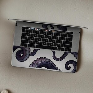 Op de afbeelding: Een laptop met een wit en paars octopus tentakel ontwerp op het toetsenbord en touchpad. De tentakels zijn diep paars met lichtere paarse zuignappen, tegen een witte achtergrond. De laptop is open op een neutrale ondergrond.