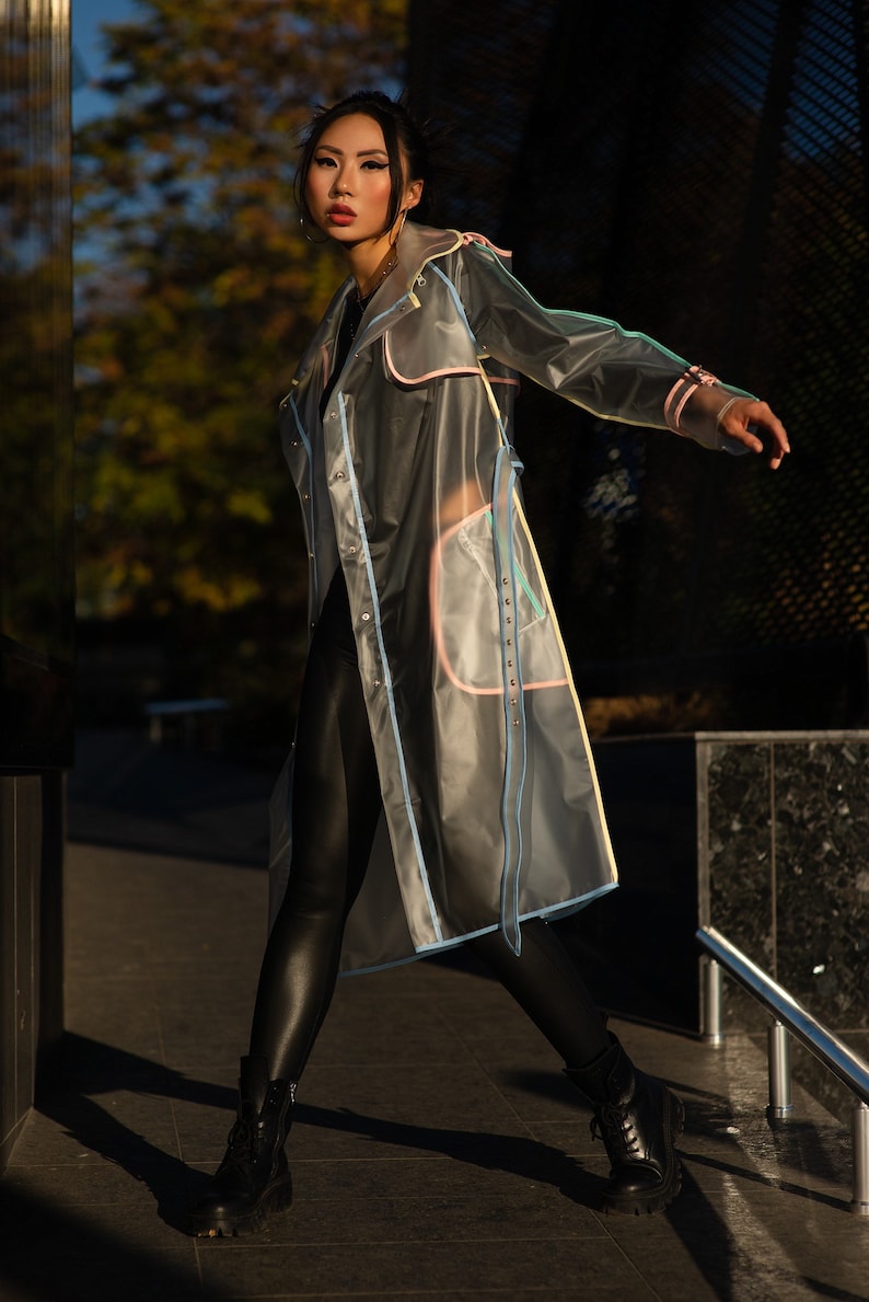 May include: A woman wearing a transparent raincoat with colorful trim and black leggings. She is standing on a sidewalk with a metal railing in the background.