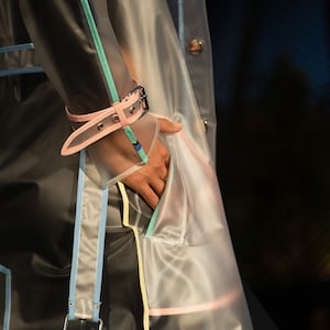 May include: A transparent raincoat with pink, blue, and yellow trim. The raincoat has a belt and a pocket on the front.