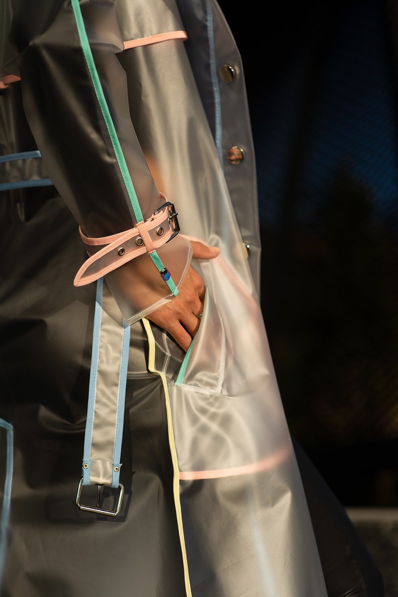 May include: A transparent raincoat with pink, blue, and yellow trim. The raincoat has a belt and a pocket on the front.