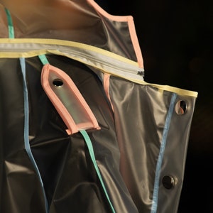 May include: A clear plastic jacket with pink, yellow, blue, and green trim. The jacket has a zipper and a pocket with a snap closure.