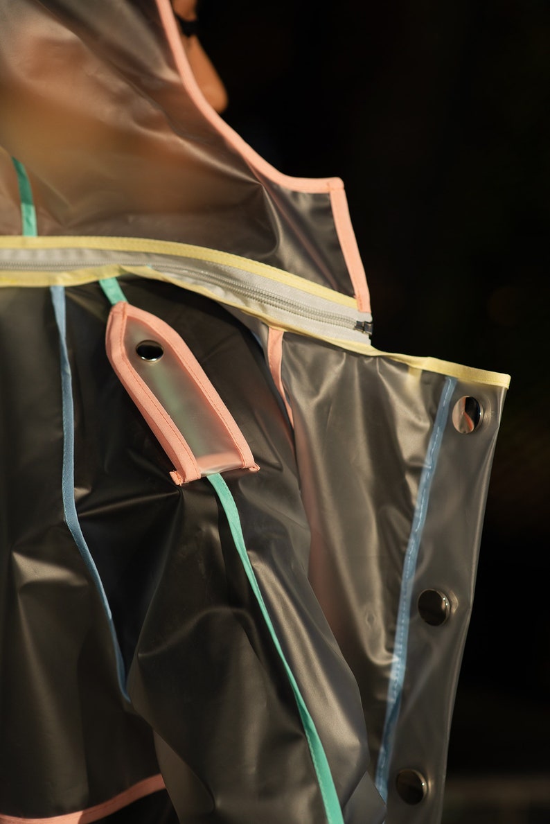 May include: A clear plastic jacket with pink, yellow, blue, and green trim. The jacket has a zipper and a pocket with a snap closure.