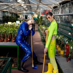 Two people in a greenhouse, one wearing a blue jumpsuit and the other wearing a neon green dress, both holding brooms.