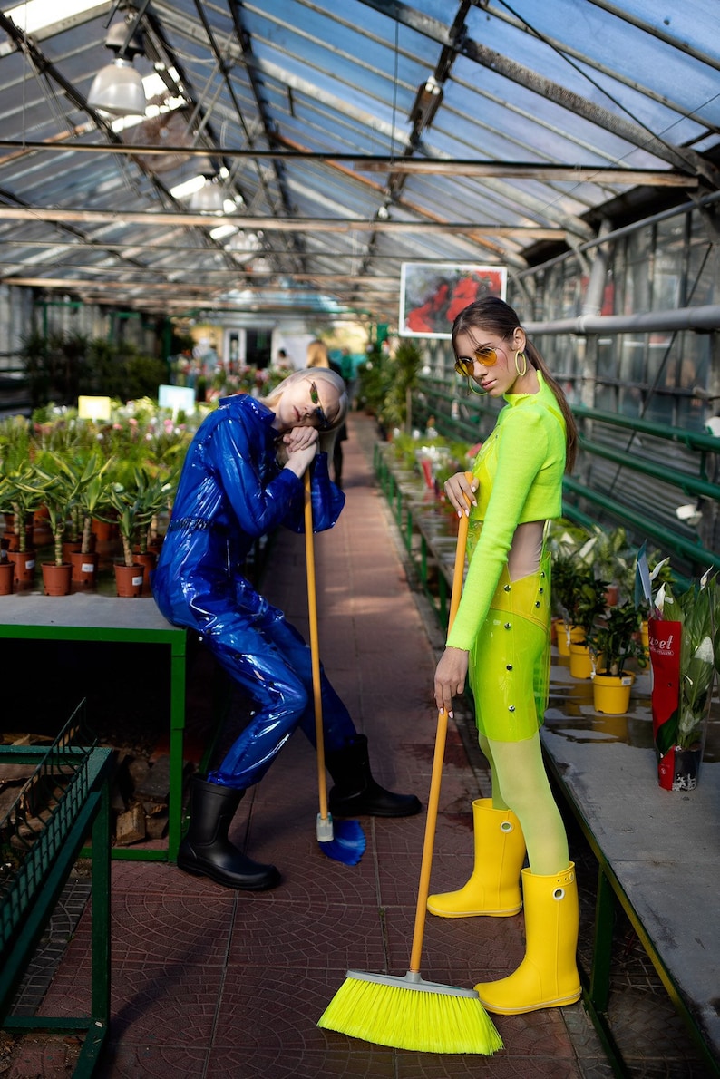 Two people in a greenhouse, one wearing a blue jumpsuit and the other wearing a neon green dress, both holding brooms.