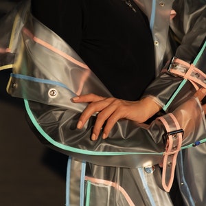 May include: A clear plastic raincoat with colorful trim. The raincoat has a light blue, pink, and yellow trim. The raincoat has a belt and a snap closure.