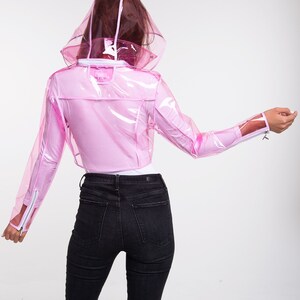 May include: A pink transparent raincoat with a hood. The raincoat is cropped and has a zipper closure. The model is wearing black jeans.