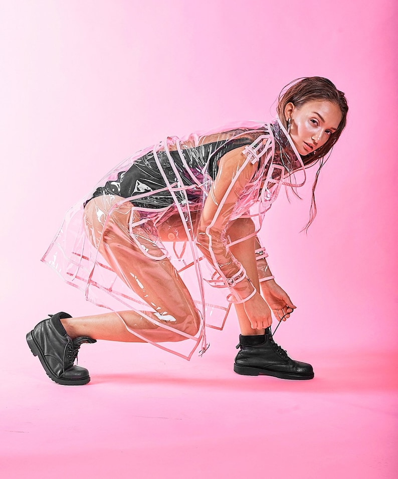 May include: A woman wearing a transparent pink raincoat with white trim, black clothing, and black lace-up boots. She is tying the laces of her boots while standing on a pink background.