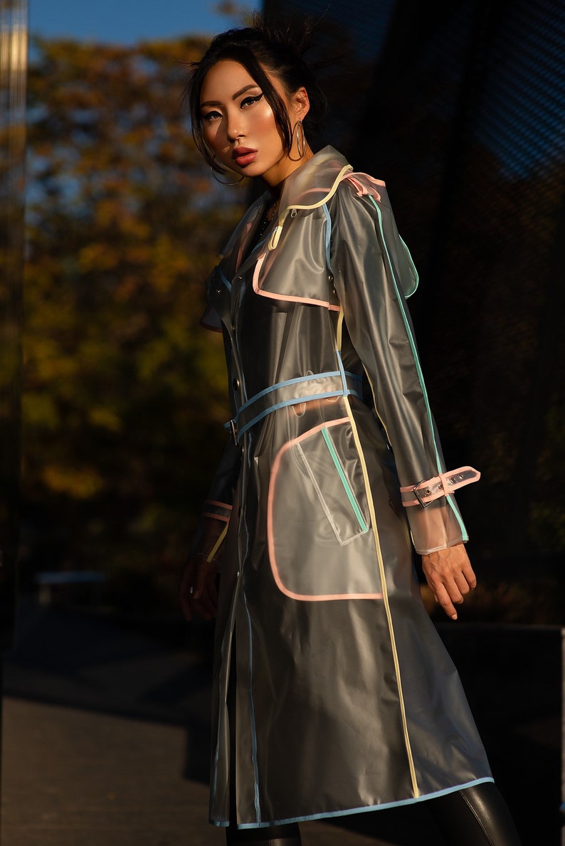 May include: A transparent raincoat with colorful trim. The raincoat has a belt and a hood. The model is wearing the raincoat and looking at the camera.