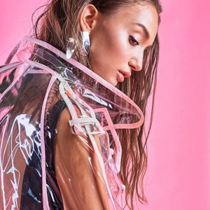 May include: A woman with long brown hair is wearing a clear plastic raincoat with pink trim. The raincoat has a hood and is see-through. The woman is looking down and her hair is covering her face. The background is a solid pink color. The text "@Domdrich&hearts;" is at the bottom of the image.
