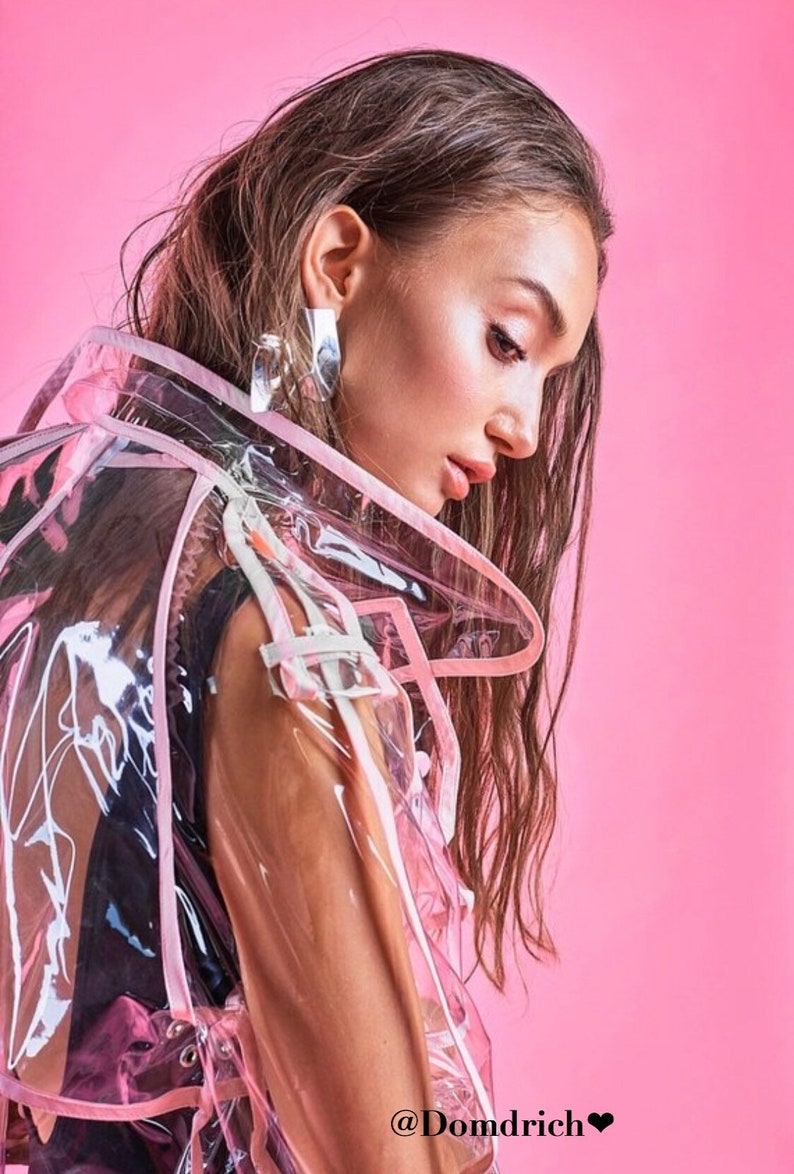 May include: A woman with long brown hair is wearing a clear plastic raincoat with pink trim. The raincoat has a hood and is see-through. The woman is looking down and her hair is covering her face. The background is a solid pink color. The text "@Domdrich&hearts;" is at the bottom of the image.