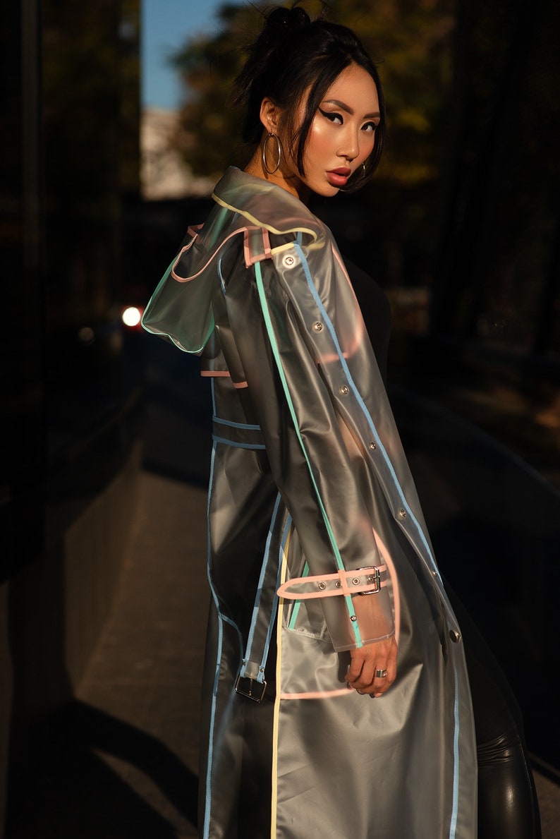 May include: A transparent raincoat with colorful trim. The raincoat has a hood and a belt. The model is wearing the raincoat and looking over their shoulder.