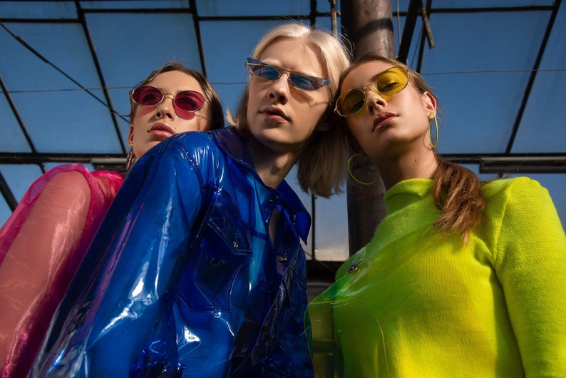 Three people wearing sunglasses, a blue jacket, a pink shirt, and a green shirt. The people are standing in front of a metal structure.