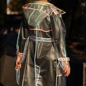 May include: A transparent raincoat with colorful trim. The raincoat has a hood and a belt. The raincoat is made of a clear plastic material and has a light blue, pink, and yellow trim.