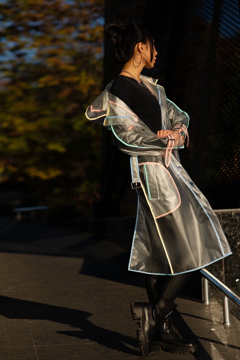 May include: A woman wearing a clear raincoat with colorful trim. The raincoat has a belt and a hood. She is wearing black pants and black boots.
