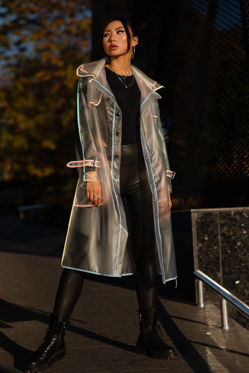 May include: A woman wearing a clear raincoat with colorful trim, black leggings, and black boots. The raincoat has a button closure and a collar.