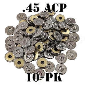 May include: A collection of silver and brass-colored metal ammunition primer caps arranged in a pile. The text .45 ACP is printed at the top and 10-PK is printed at the bottom. Each individual circular component features stamped markings.