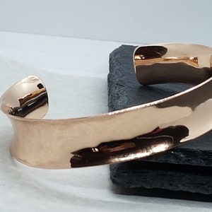 May include: A rose gold cuff bracelet with a curved, open design. The polished metal reflects light, highlighting the smooth, wide band. The bracelet sits on a dark, textured surface, showcasing its elegant simplicity. A stylish piece of jewellery.