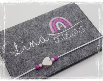 Può includere: Scatola portaoggetti in feltro grigio con un arcobaleno ricamato in bianco, il nome "Lina" e la data "05.06.2023". Un ciondolo a forma di cuore bianco con perline rosa è attaccato alla scatola.